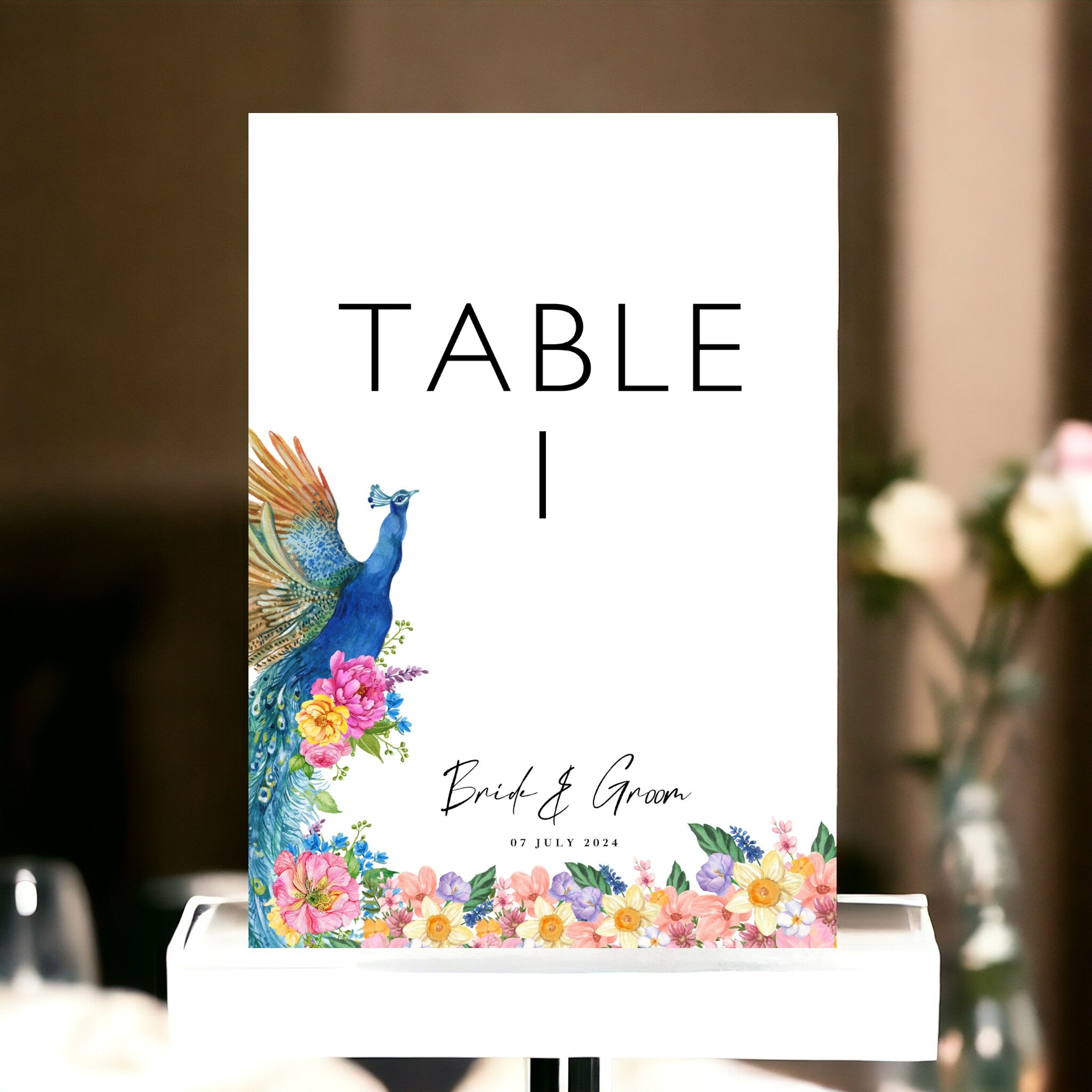 Personalised Indian wedding table number card printed double sided on 350gsm silk card. Clean white design with vibrant illustrated peacock, colourful wildflowers and botanicals, bold table number, custom couple names and wedding date. Shown in wire stand on wedding table with pink roses. By Krafty Kaur.