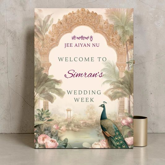 Personalised Punjabi wedding week welcome sign printed on 3mm foamex. Blush and sage Mughal arch with ornate gold carving, peacock, tropical palms, pink peonies and misty Indian garden illustration. Jee Aiyan Nu in Gurmukhi, custom name in plum script. A1 or A2. By Krafty Kaur.