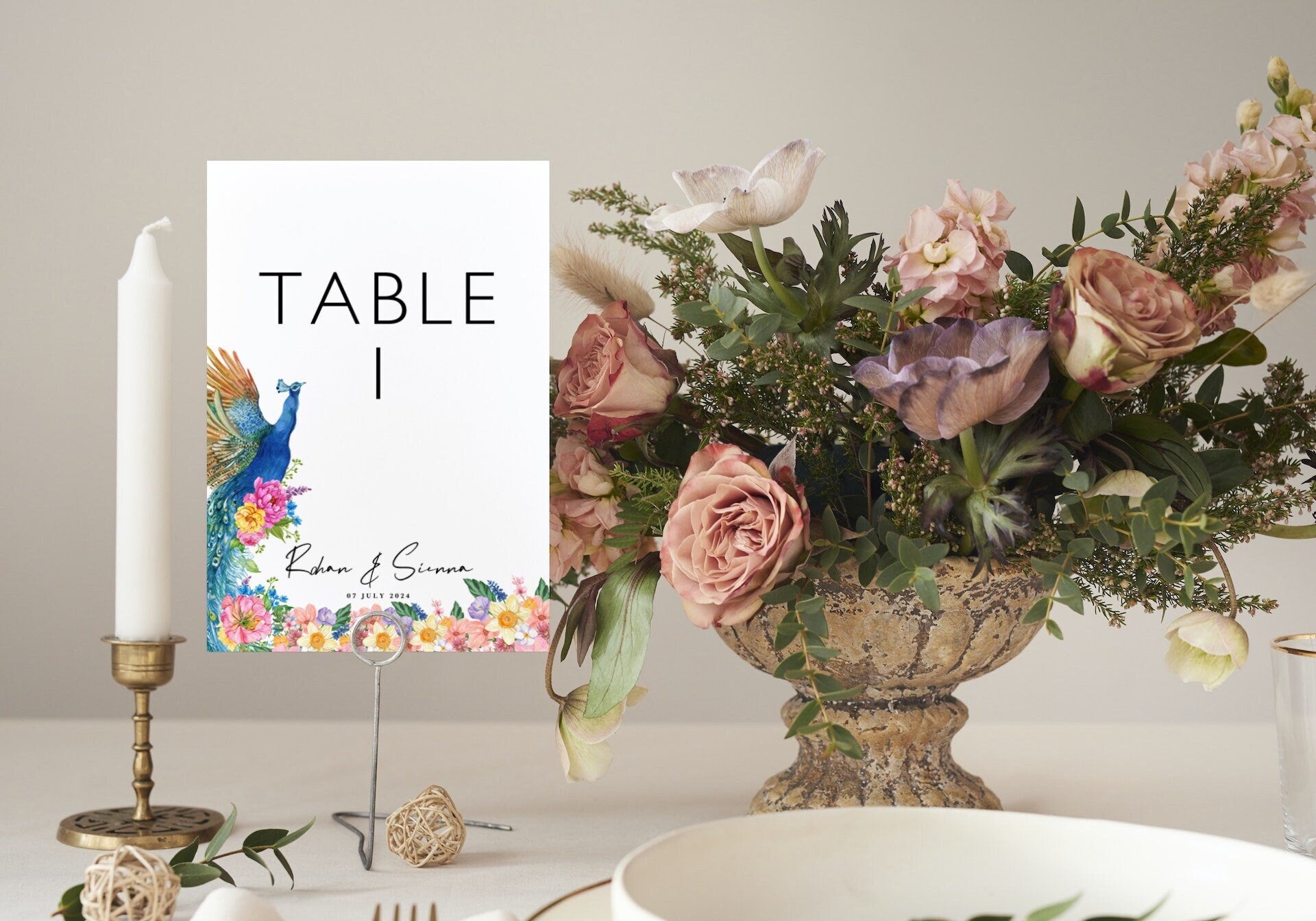 Custom peacock Indian wedding table number on wire stand beside candlestick and blush flower arrangement. White card with colourful peacock and wildflowers, personalised couple names and date. 350gsm silk. Krafty Kaur.