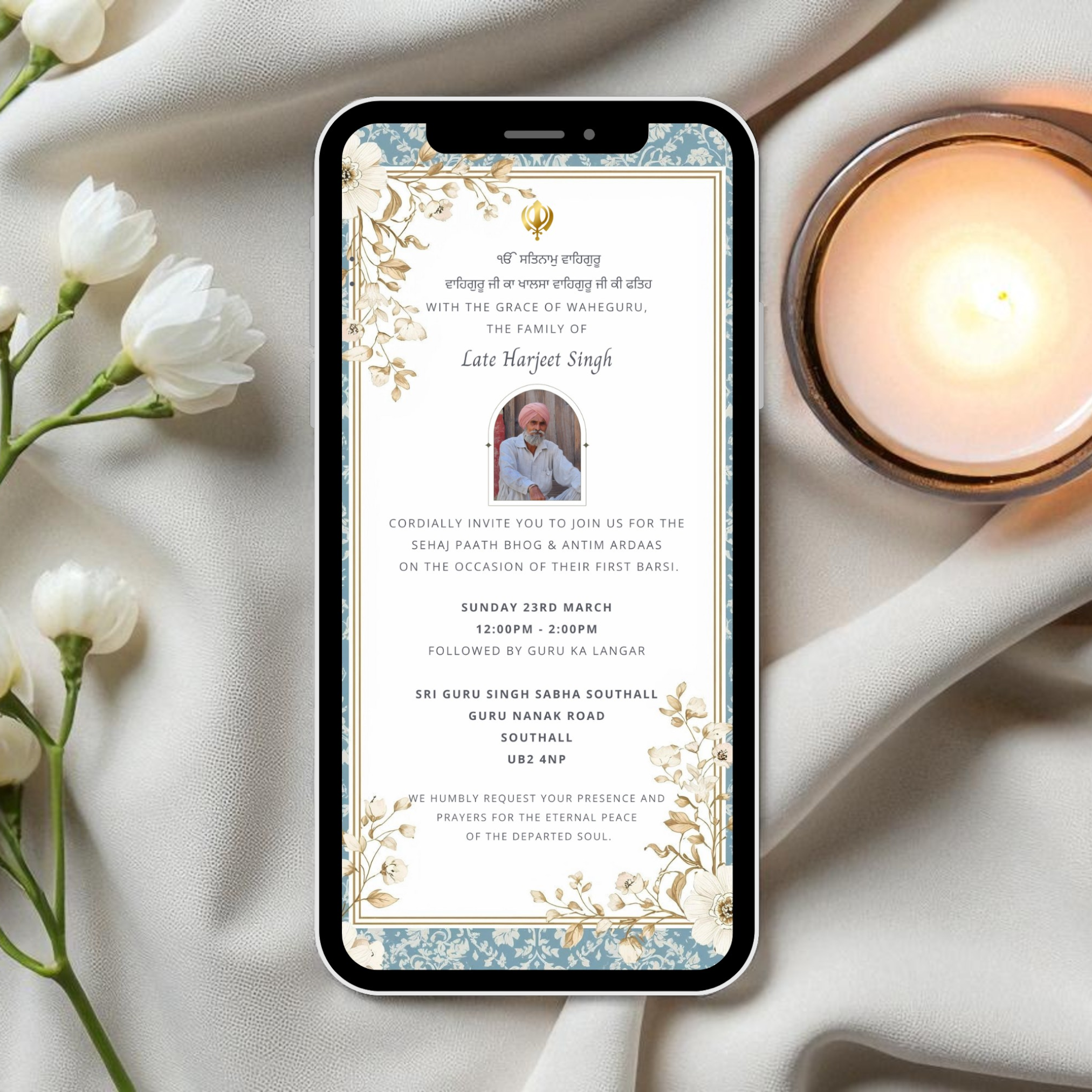 Phone displaying a digital sikh death anniversary barsi invitation with flowers and a candle on a soft fabric background