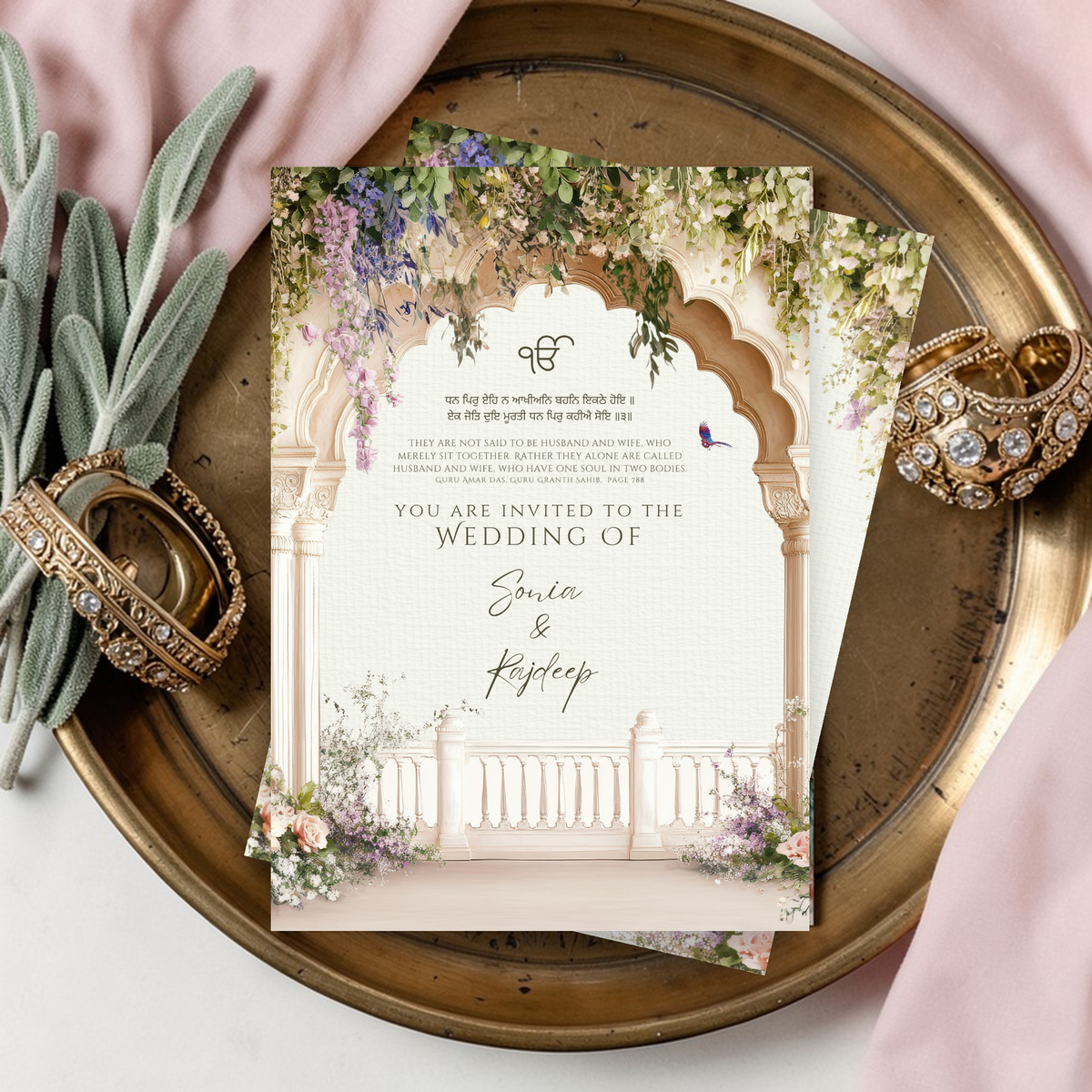 Sikh wedding invitation suite flat lay on a brass tray with choora bangles and sage stems, showing cover page with Ik Onkar symbol, Guru Granth Sahib shabad in Gurmukhi and English, and wisteria floral Mughal arch design personalised with names Sonia and Rajdeep