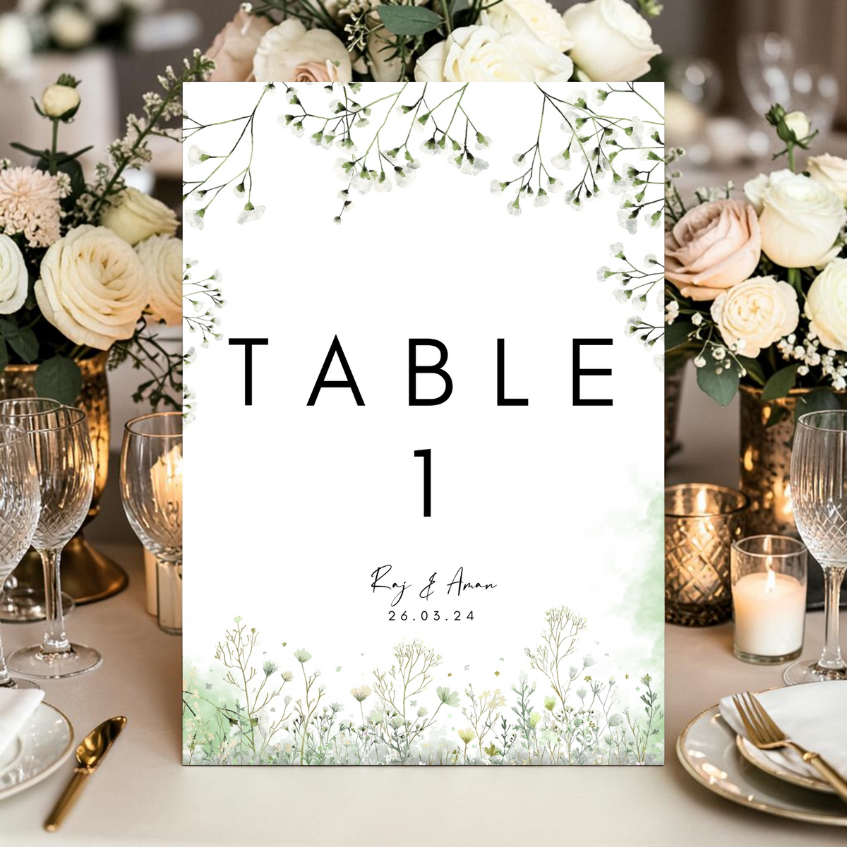 Personalised wedding table number card printed double sided on A5 350gsm silk card. Clean white design with watercolour gypsophila and sage green botanical wildflowers, bold modern table number, custom couple names and wedding date in script. Shown in gold stand on reception table with cream roses. By Krafty Kaur.
