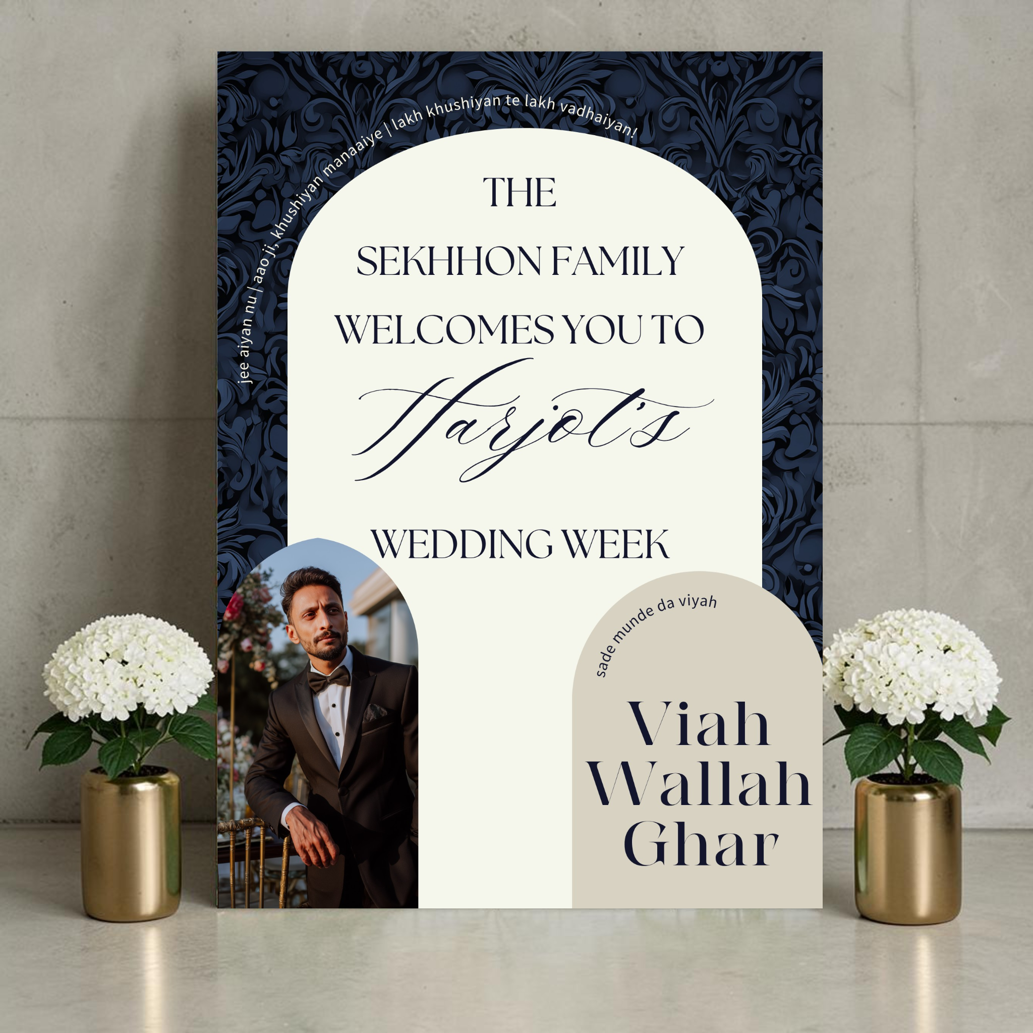 Punjabi wedding week welcome sign on a wooden easel beside white hydrangeas in a brass vase, featuring a groom photo in an arch cutout on a bold navy damask background with cream arch panel reading "The Sekhhon Family Welcomes You to Harjot's Wedding Week" with Viah Wallah Ghar and Sade Munde Da Viyah text
