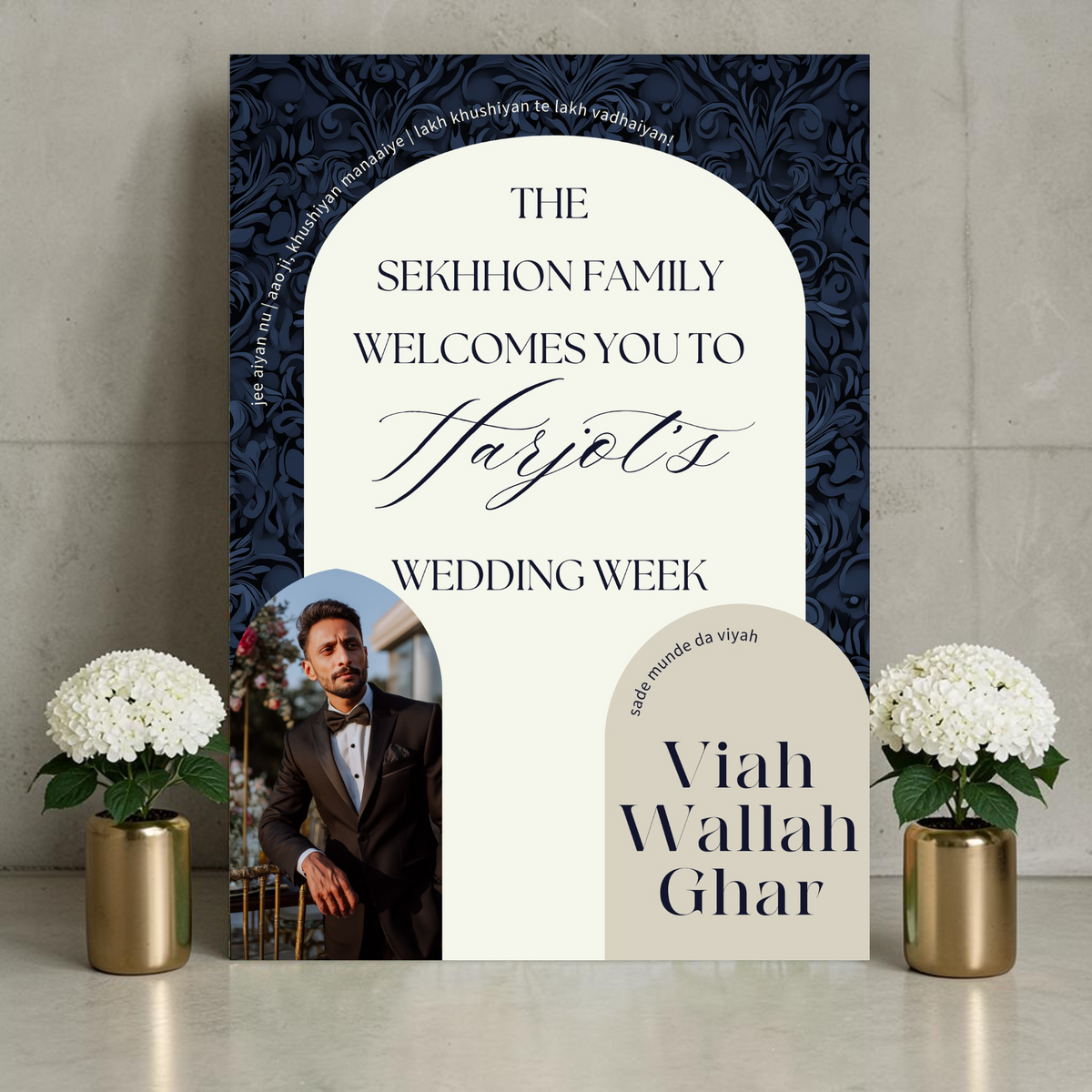 Punjabi wedding week welcome sign on a wooden easel beside white hydrangeas in a brass vase, featuring a groom photo in an arch cutout on a bold navy damask background with cream arch panel reading "The Sekhhon Family Welcomes You to Harjot's Wedding Week" with Viah Wallah Ghar and Sade Munde Da Viyah text
